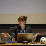 Woman in a blazer speaks at a panel desk with a laptop and microphone, bike helmet on the desk nearby, OSCHRIN nameplate visible.