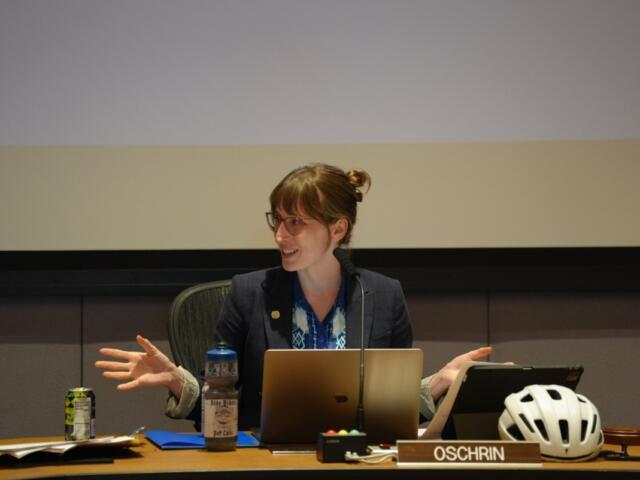 Woman in a blazer speaks at a panel desk with a laptop and microphone, bike helmet on the desk nearby, OSCHRIN nameplate visible.