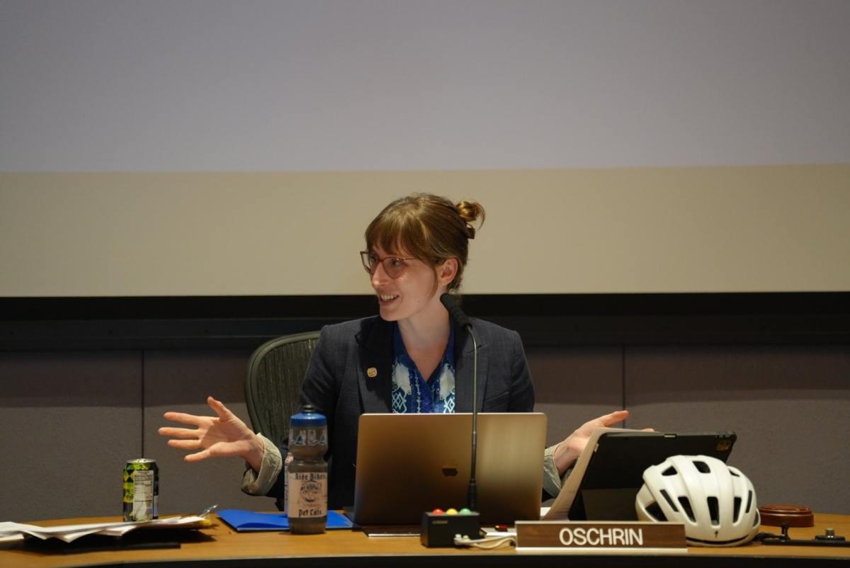 Woman in a blazer speaks at a panel desk with a laptop and microphone, bike helmet on the desk nearby, OSCHRIN nameplate visible.