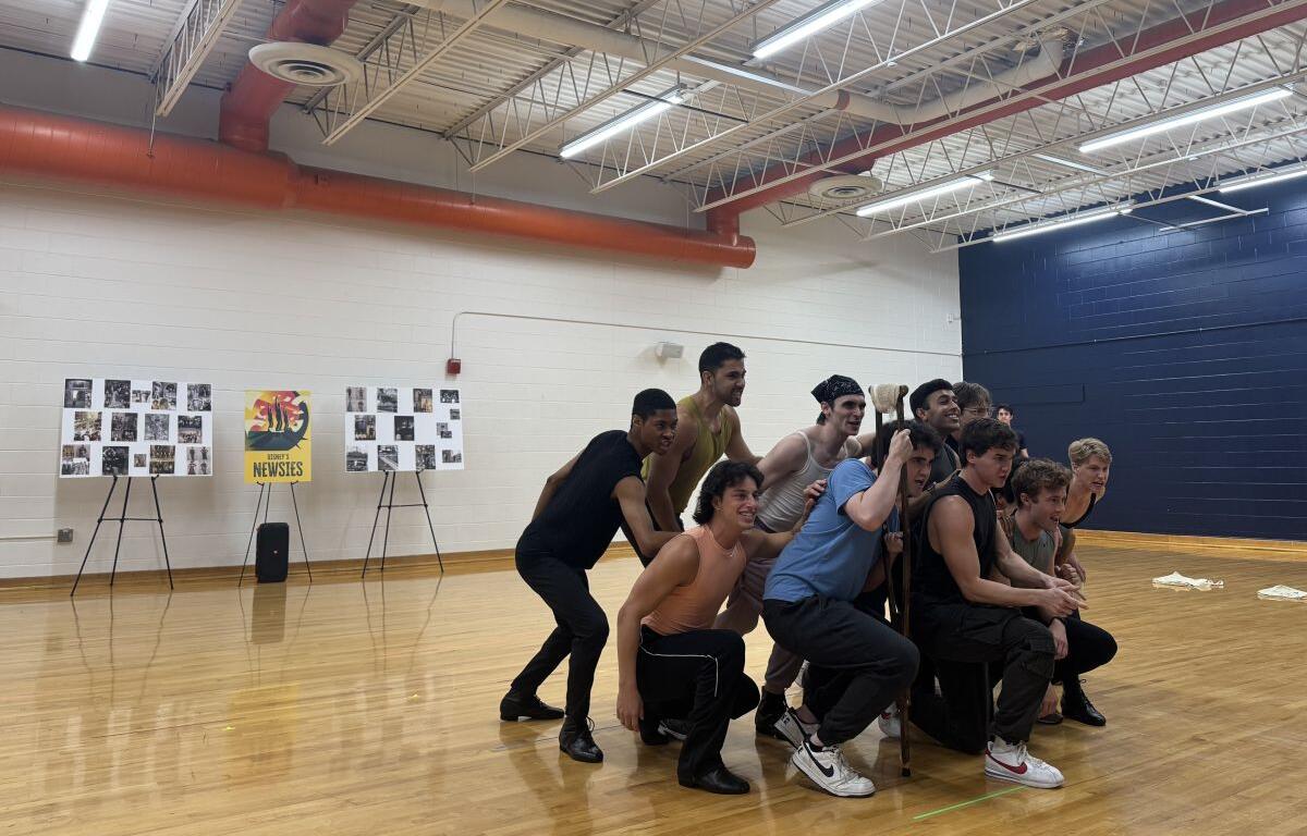 Group of young men posing in a gym: some crouching, one standing with a staff, smiles on their faces against a wooden floor and blue wall backdrop.