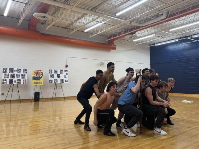 Group of young men posing in a gym: some crouching, one standing with a staff, smiles on their faces against a wooden floor and blue wall backdrop.