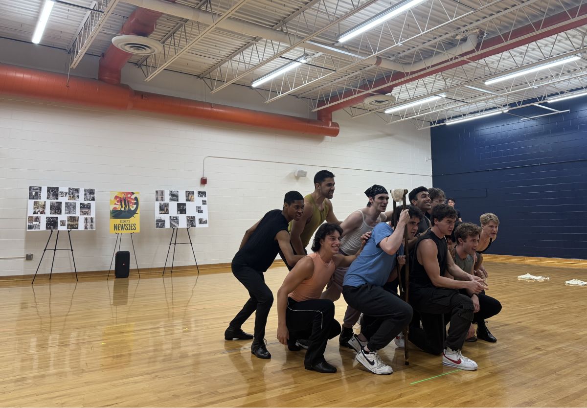 Group of young men posing in a gym: some crouching, one standing with a staff, smiles on their faces against a wooden floor and blue wall backdrop.
