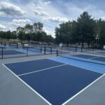 Outdoor blue tennis courts with nets, surrounded by black chain-link fencing and trees under a partly cloudy sky.