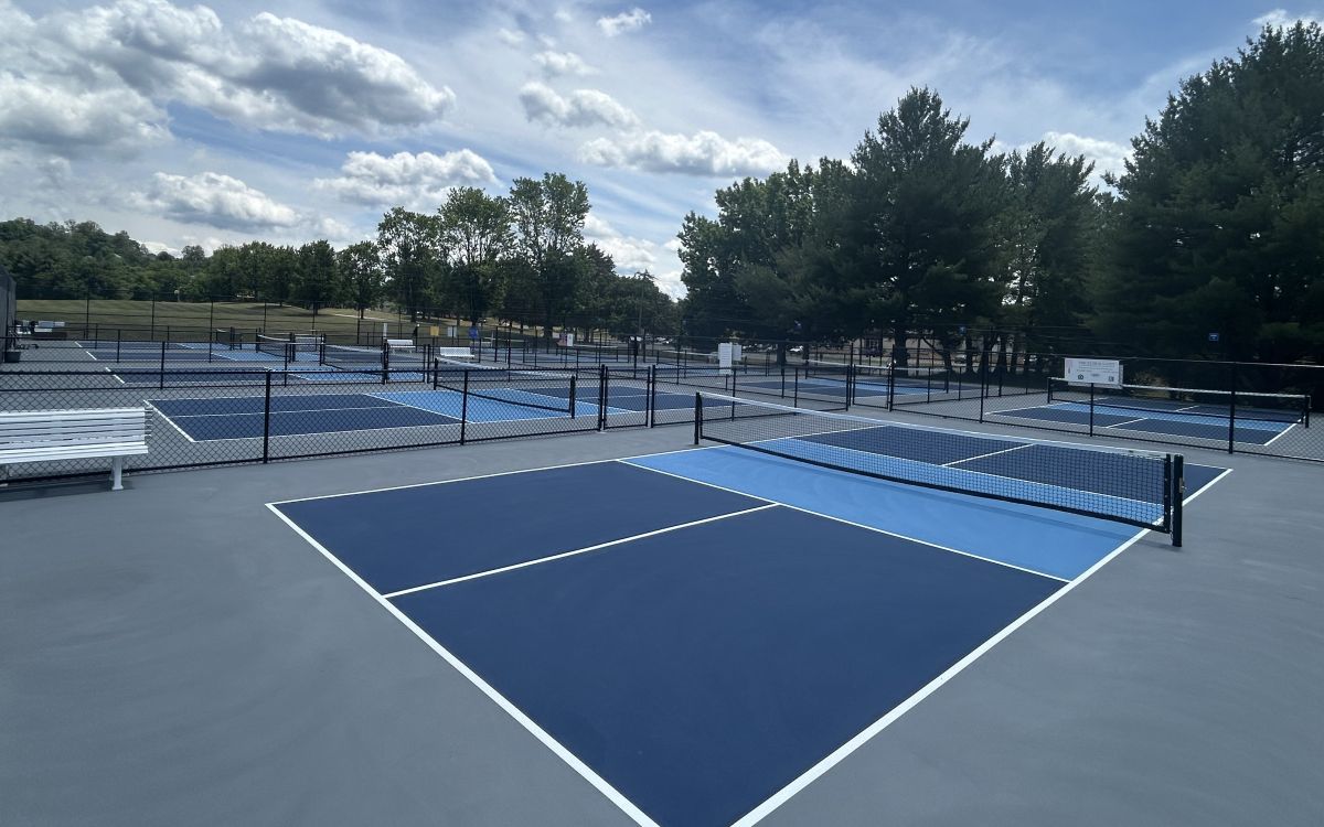Outdoor blue tennis courts with nets, surrounded by black chain-link fencing and trees under a partly cloudy sky.