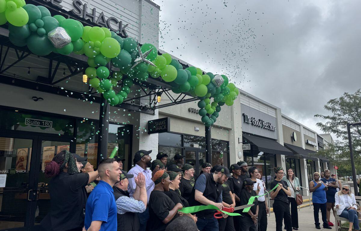 Ribbon cutting ceremony outside Shake Shack with a green balloon arch, confetti, and cheering attendees.