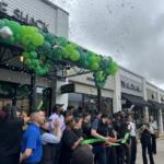 Ribbon cutting ceremony outside Shake Shack with a green balloon arch, confetti, and cheering attendees.