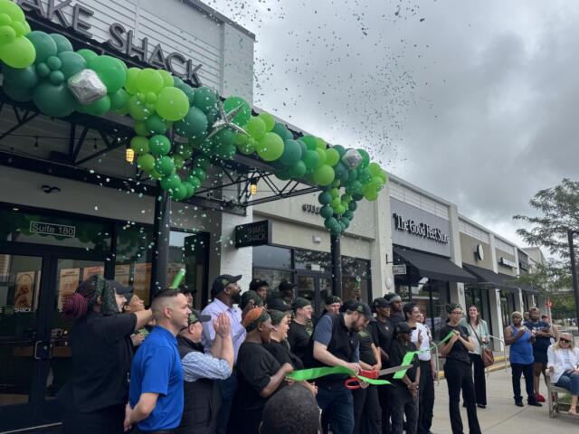 Ribbon cutting ceremony outside Shake Shack with a green balloon arch, confetti, and cheering attendees.