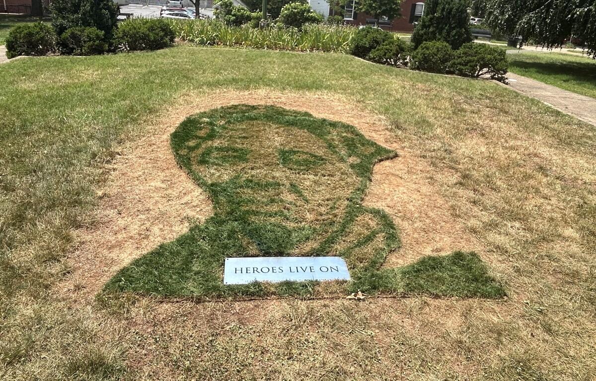 Grass mosaic of a person's silhouette on a small hill, with a plaque reading 'HEROES LIVE ON' at the bottom.