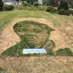 Grass mosaic of a person's silhouette on a small hill, with a plaque reading 'HEROES LIVE ON' at the bottom.