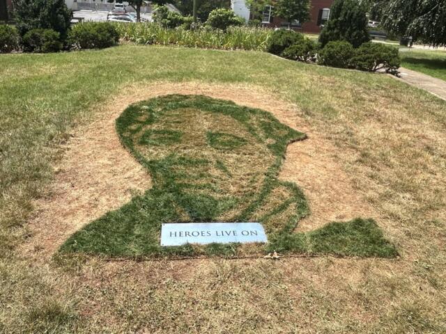 Grass mosaic of a person's silhouette on a small hill, with a plaque reading 'HEROES LIVE ON' at the bottom.