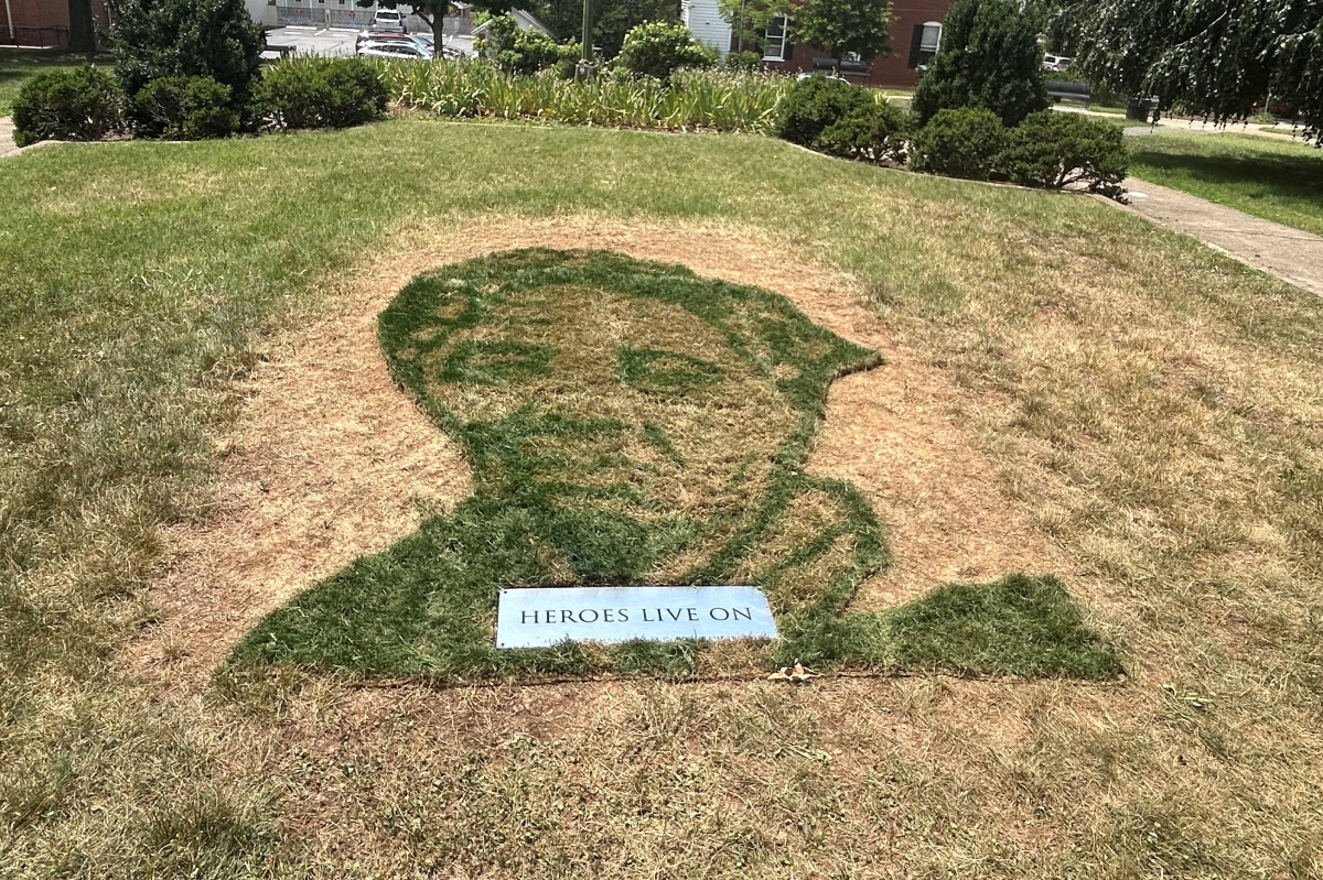 Grass mosaic of a person's silhouette on a small hill, with a plaque reading 'HEROES LIVE ON' at the bottom.