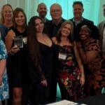 Group of smiling professionals posing at an awards ceremony; several hold plaques with a teal backdrop behind them.