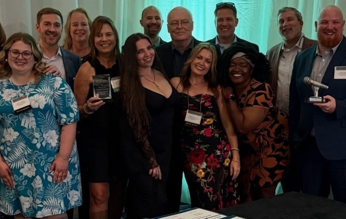 Group of smiling professionals posing at an awards ceremony; several hold plaques with a teal backdrop behind them.