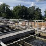 Industrial water treatment plant with metal catwalks, railings, and a red lifebuoy, trees in the background on a sunny day.