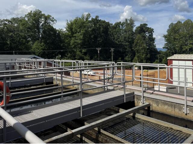 Industrial water treatment plant with metal catwalks, railings, and a red lifebuoy, trees in the background on a sunny day.