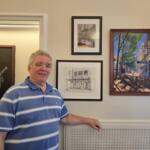 Smiling man in a blue striped polo standing beside framed artworks on a gallery wall.