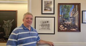 Smiling man in a blue striped polo standing beside framed artworks on a gallery wall.