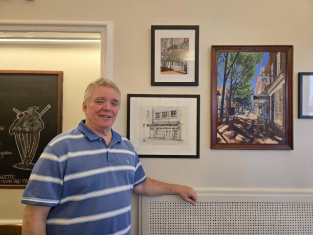 Smiling man in a blue striped polo standing beside framed artworks on a gallery wall.