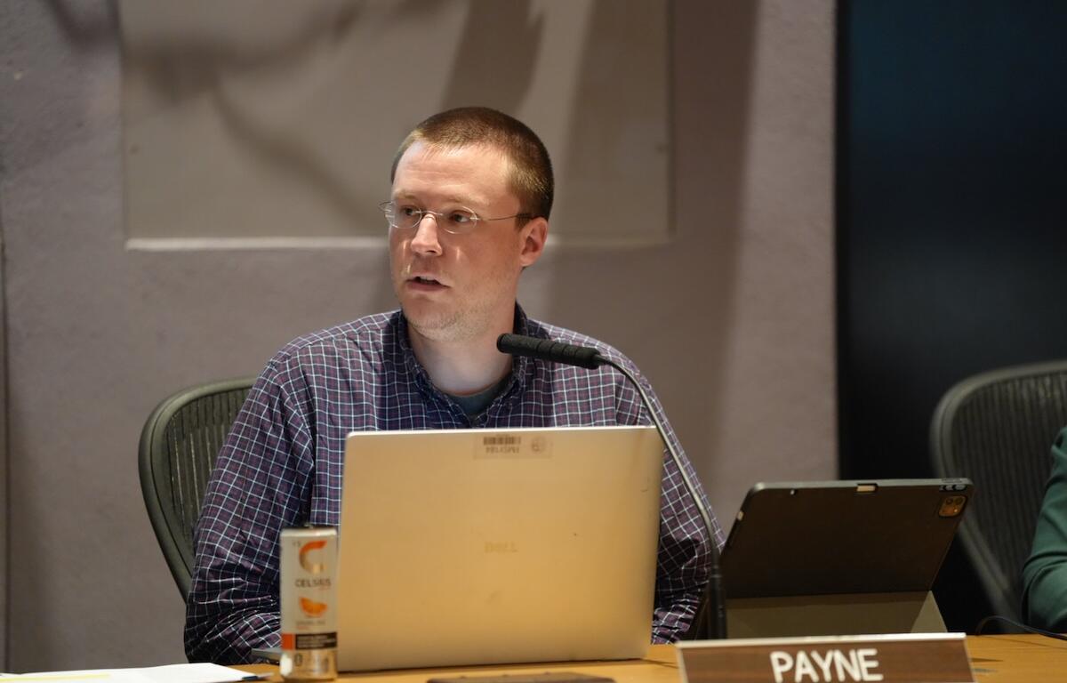 Man seated at a conference table with a laptop, microphone, and a tablet, looking to the side; nameplate reads PAYNE in front of him