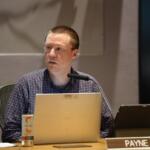 Man seated at a conference table with a laptop, microphone, and a tablet, looking to the side; nameplate reads PAYNE in front of him