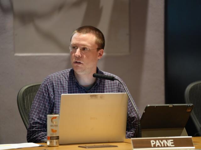 Man seated at a conference table with a laptop, microphone, and a tablet, looking to the side; nameplate reads PAYNE in front of him