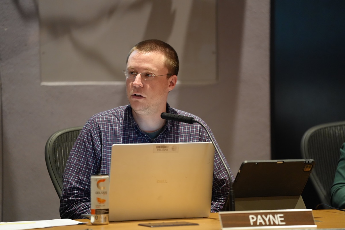 Man seated at a conference table with a laptop, microphone, and a tablet, looking to the side; nameplate reads PAYNE in front of him