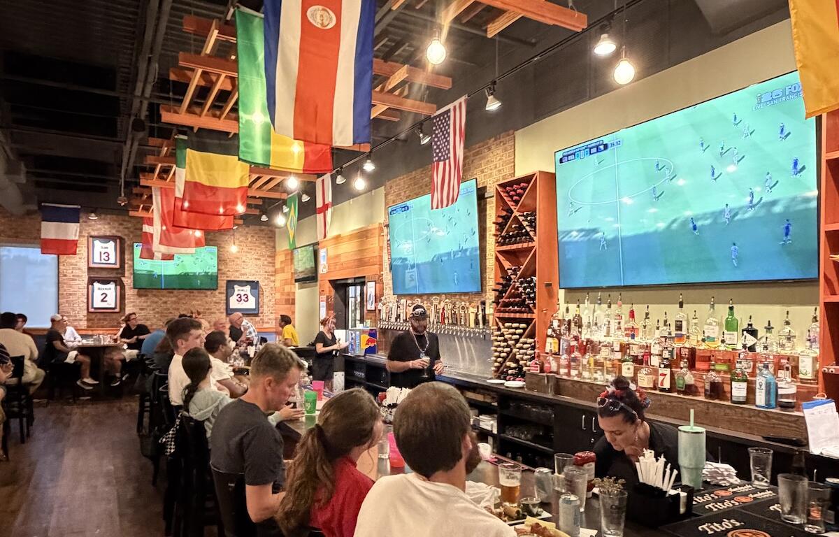 Sports bar interior with flags hanging from the ceiling, a long bar on the right, and patrons seated at tables watching a soccer match on two large wall-mounted TVs.