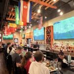 Sports bar interior with flags hanging from the ceiling, a long bar on the right, and patrons seated at tables watching a soccer match on two large wall-mounted TVs.