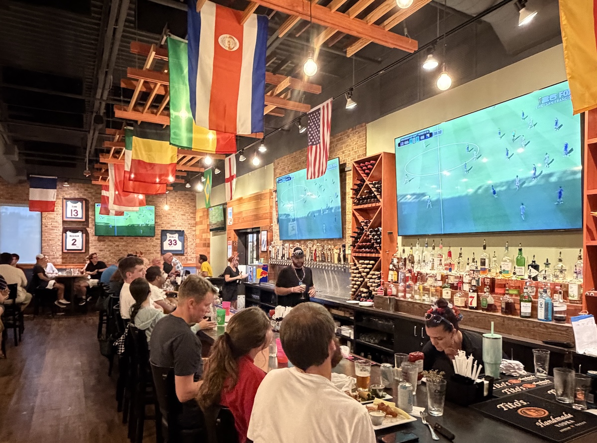 Sports bar interior with flags hanging from the ceiling, a long bar on the right, and patrons seated at tables watching a soccer match on two large wall-mounted TVs.
