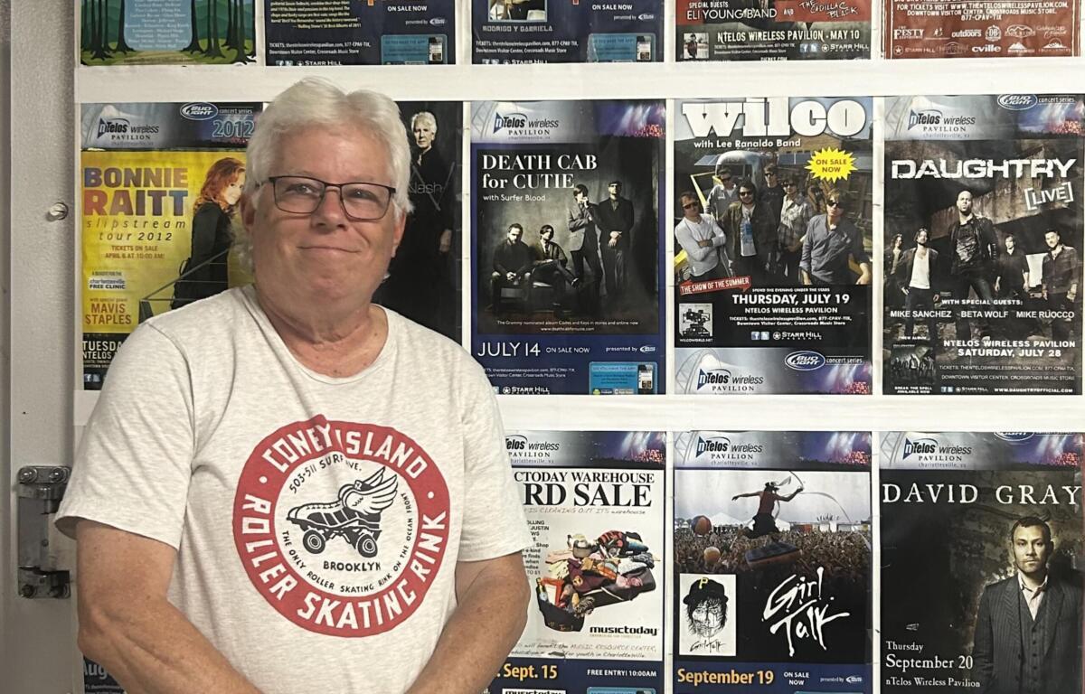 Older man with gray hair and glasses standing in front of a wall covered with colorful concert posters.