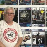 Older man with gray hair and glasses standing in front of a wall covered with colorful concert posters.