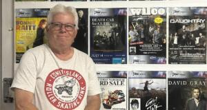 Older man with gray hair and glasses standing in front of a wall covered with colorful concert posters.
