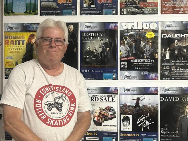 Older man with gray hair and glasses standing in front of a wall covered with colorful concert posters.