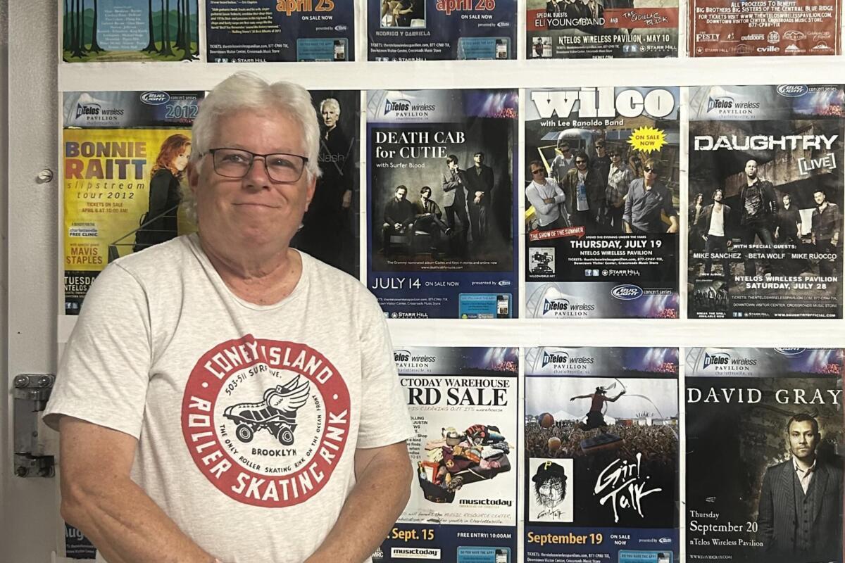 Older man with gray hair and glasses standing in front of a wall covered with colorful concert posters.