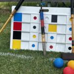 Giant outdoor Connect Four-style board on grass with colorful balls nearby and wooden mallets resting against it.