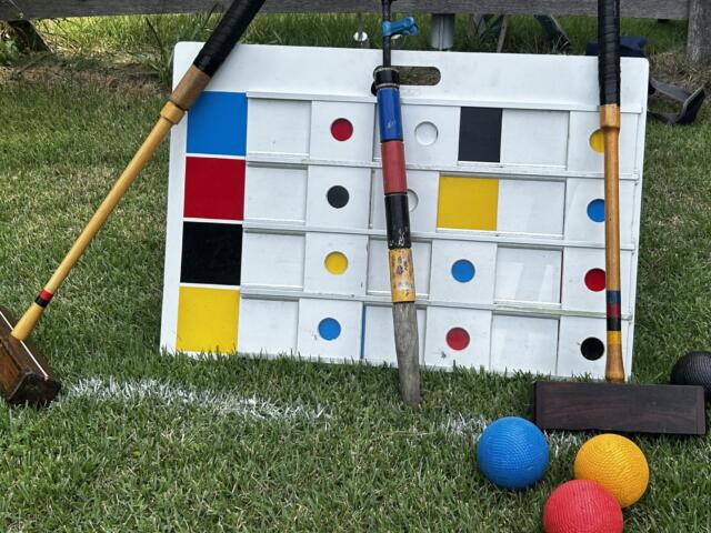 Giant outdoor Connect Four-style board on grass with colorful balls nearby and wooden mallets resting against it.