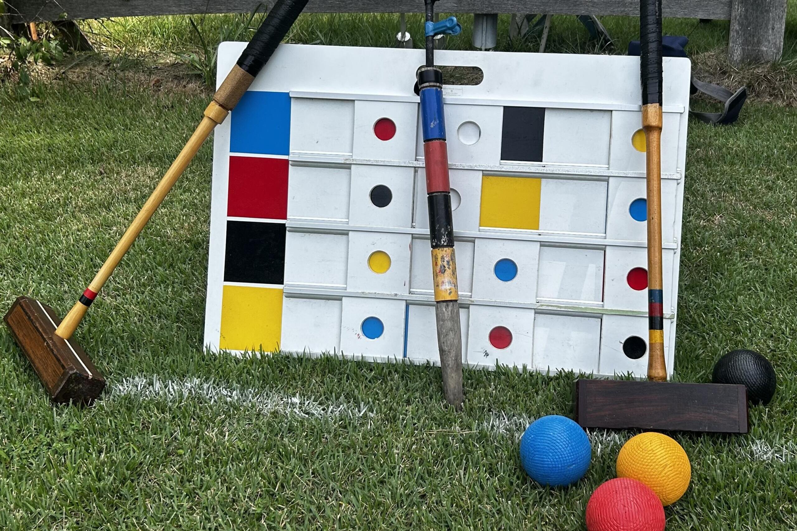 Giant outdoor Connect Four-style board on grass with colorful balls nearby and wooden mallets resting against it.