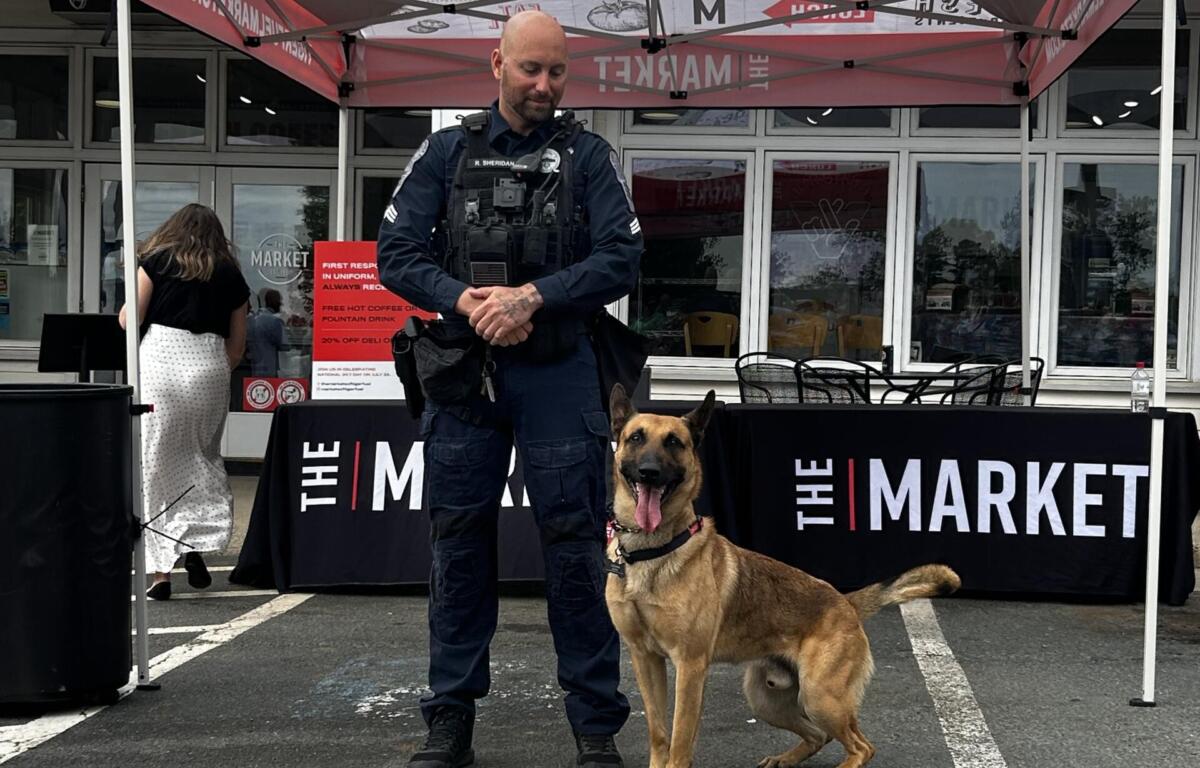 Police officer in tactical vest standing with a German Shepherd at an event under a red canopy outside a market venue.