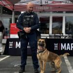 Police officer in tactical vest standing with a German Shepherd at an event under a red canopy outside a market venue.
