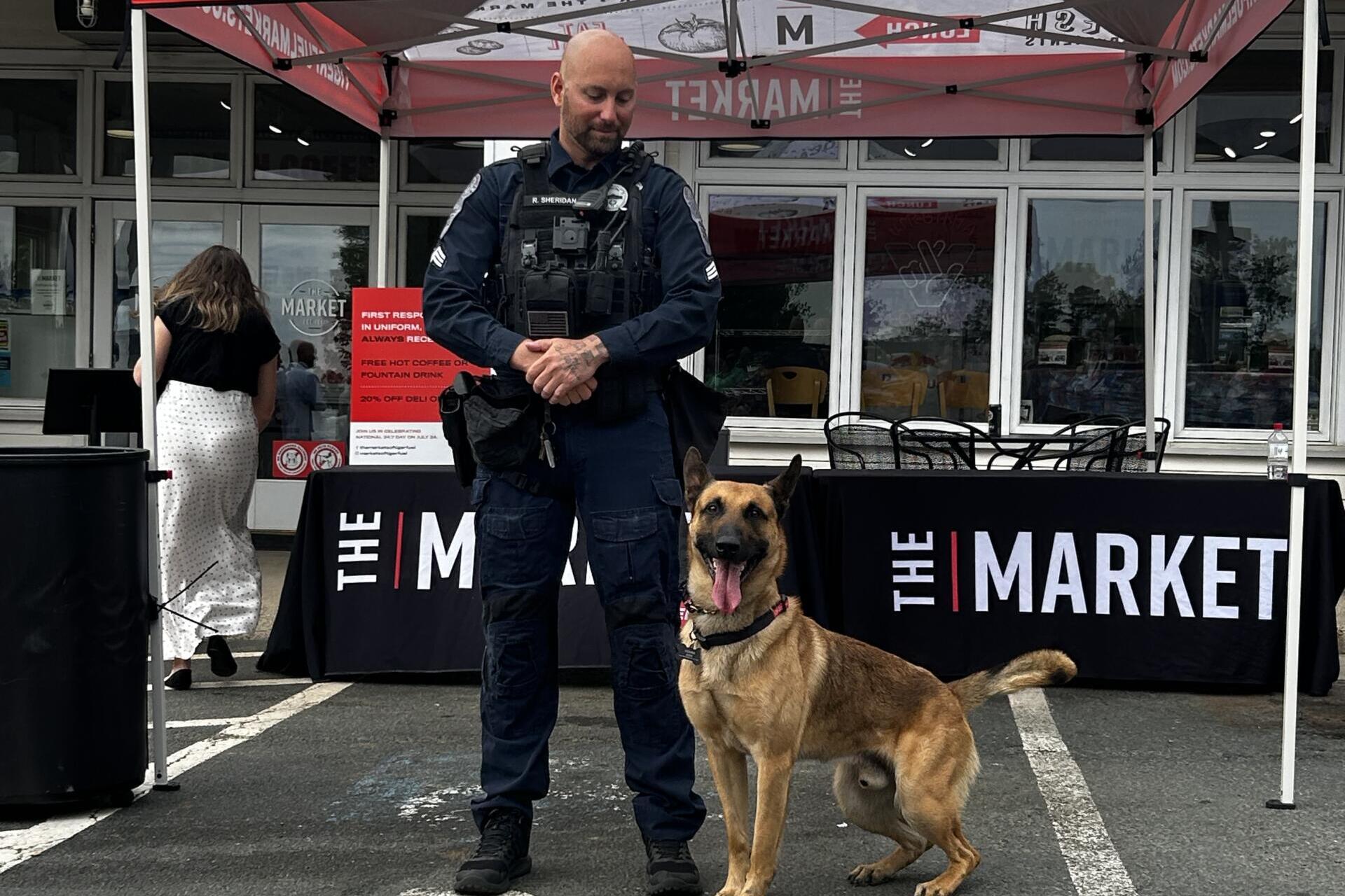 Police officer in tactical vest standing with a German Shepherd at an event under a red canopy outside a market venue.