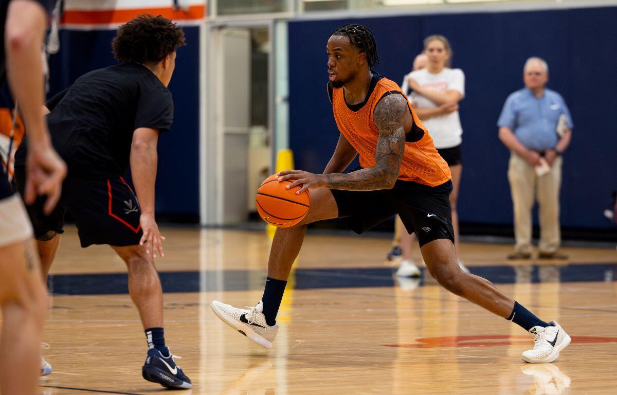 Basketball drill: a player in an orange jersey dribbles the ball past a defender on an indoor court, gym in background, spectators blurred.