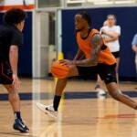 Basketball drill: a player in an orange jersey dribbles the ball past a defender on an indoor court, gym in background, spectators blurred.