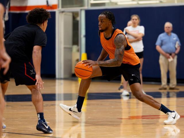 Basketball drill: a player in an orange jersey dribbles the ball past a defender on an indoor court, gym in background, spectators blurred.