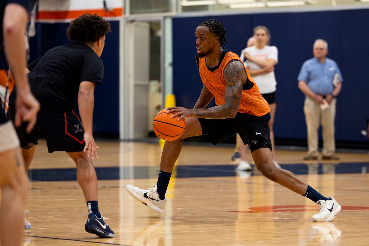 Basketball drill: a player in an orange jersey dribbles the ball past a defender on an indoor court, gym in background, spectators blurred.