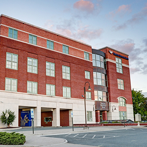 Exterior view of a red brick, four-story building with white trim and a curved glass corner entry along a street.
