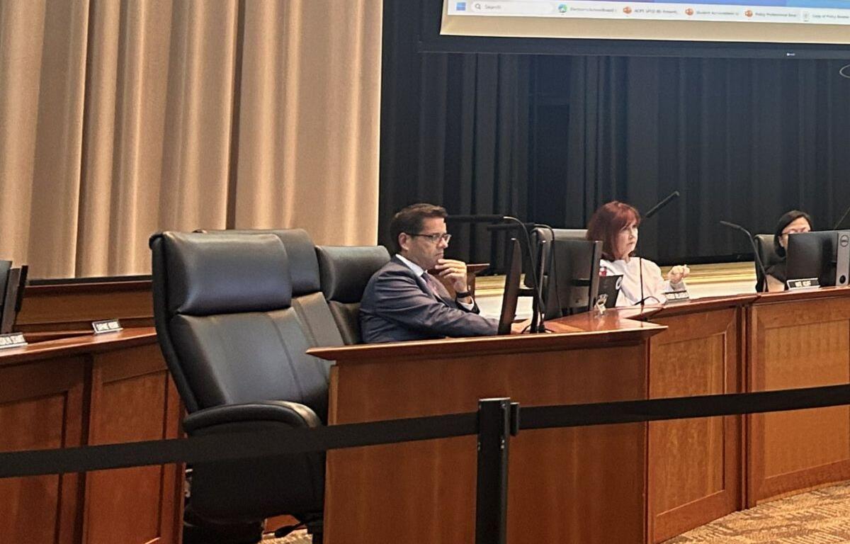 Three panelists seated at a curved wooden desk in a formal meeting room, each with a monitor in front of them.