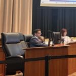 Three panelists seated at a curved wooden desk in a formal meeting room, each with a monitor in front of them.