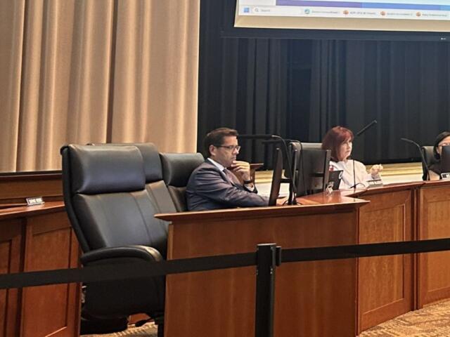 Three panelists seated at a curved wooden desk in a formal meeting room, each with a monitor in front of them.