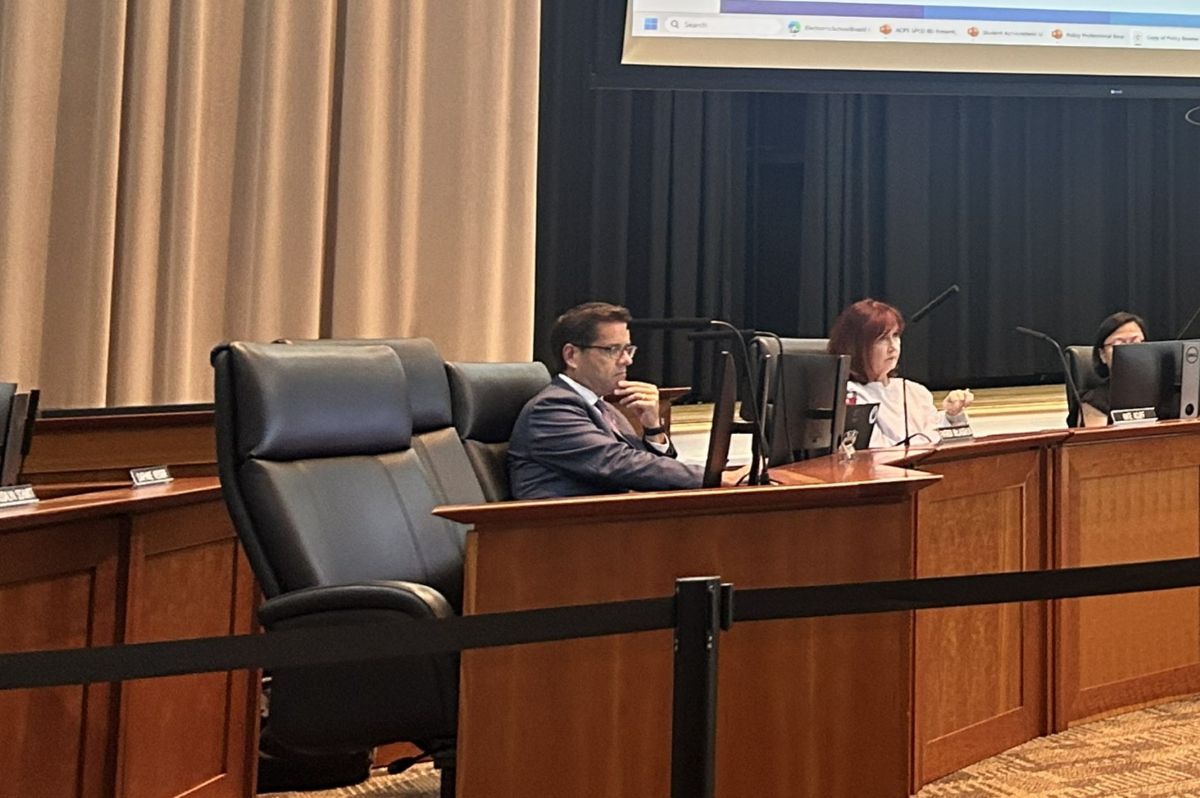 Three panelists seated at a curved wooden desk in a formal meeting room, each with a monitor in front of them.