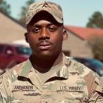 Portrait of a U.S. Army soldier in desert camouflage, cap on, with the name Anderson on the chest, standing outdoors near buildings and cars.